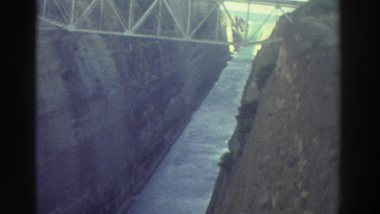 Metal truss bridge spanning a narrow rock-cut canal, photographed from near bridge level looking down at blue-purple water between steep walls.