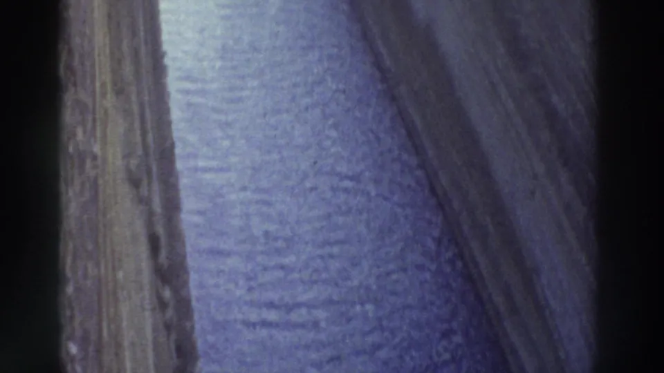 8mm film frame, Greece 1976 — close forward view along a narrow canal with steep layered rock walls and rippled blue-purple water surface