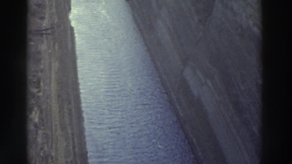 8mm film frame, Greece 1976 — forward-looking shot along a straight narrow canal with tall layered rock walls and grey-blue water