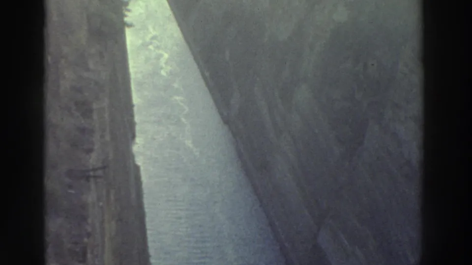 8mm film frame, Greece 1976 — wider view of a straight canal flanked by towering rock cliffs with pale blue water and sparse vegetation at the cliff tops