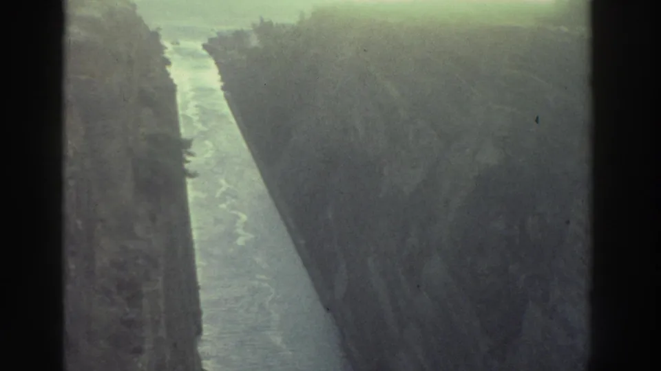 8mm film frame, Greece 1976 — straight canal between sheer rock walls, pale waterway receding toward a brighter opening, sparse scrub at cliff tops