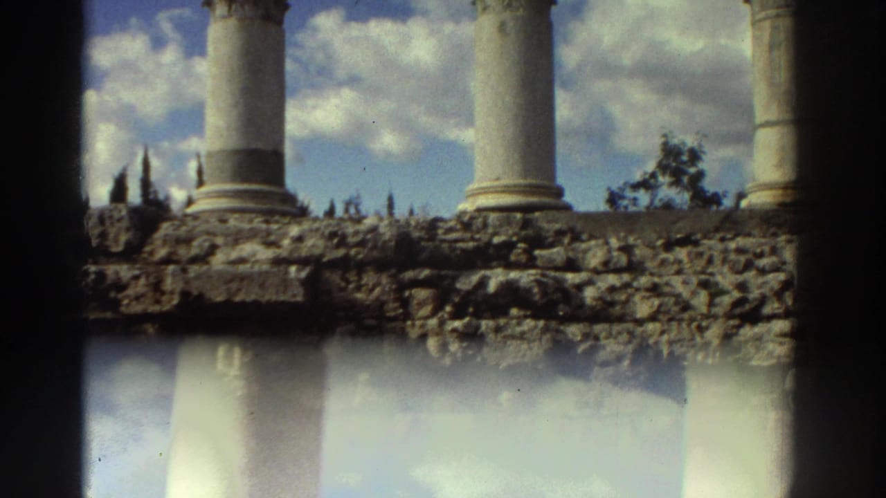 Tall Corinthian columns viewed closer with column bases and ruined stone platform below; light flare in lower frame and cypress trees in background.