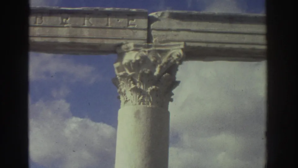 Corinthian capital and partial entablature inscription at a Greek archaeological site, 1976 8mm film; camera has panned slightly from preceding frame.
