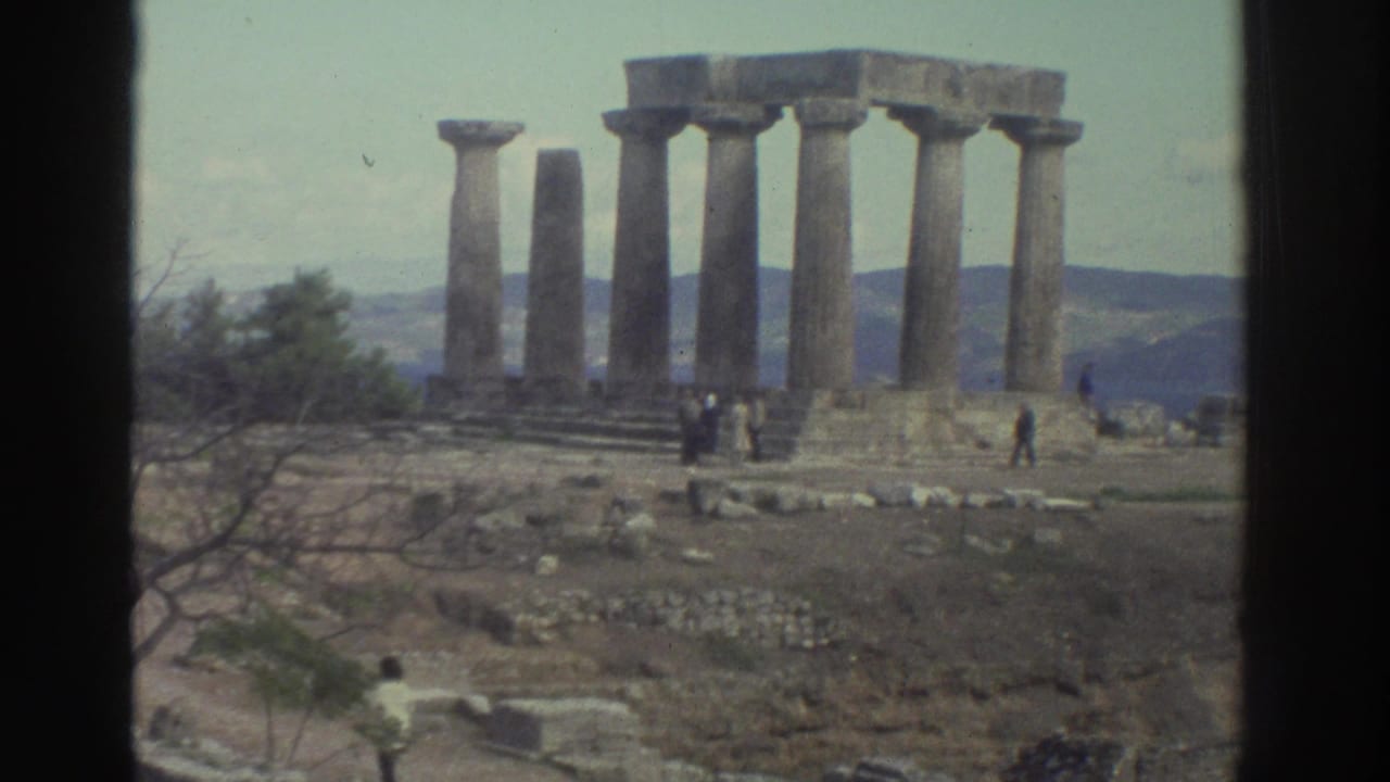 Wide 8mm shot of standing ancient Doric columns on a rocky hilltop, visitors at the base, mountains behind.
