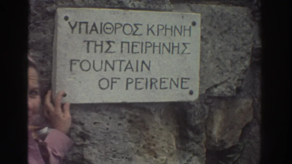 8mm close-up of a stone bilingual sign for the Fountain of Peirene (ΥΠΑΙΘΡΟΣ ΚΡΗΝΗ ΤΗΣ ΠΕΙΡΗΝΗΣ), Greece mid-1970s; woman in pink hooded jacket partially visible at lower frame.