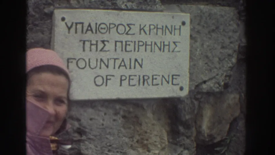 8mm frame of the Fountain of Peirene site marker, Greece mid-1970s; woman in pink hooded jacket faces camera alongside the bilingual Greek and English inscription.
