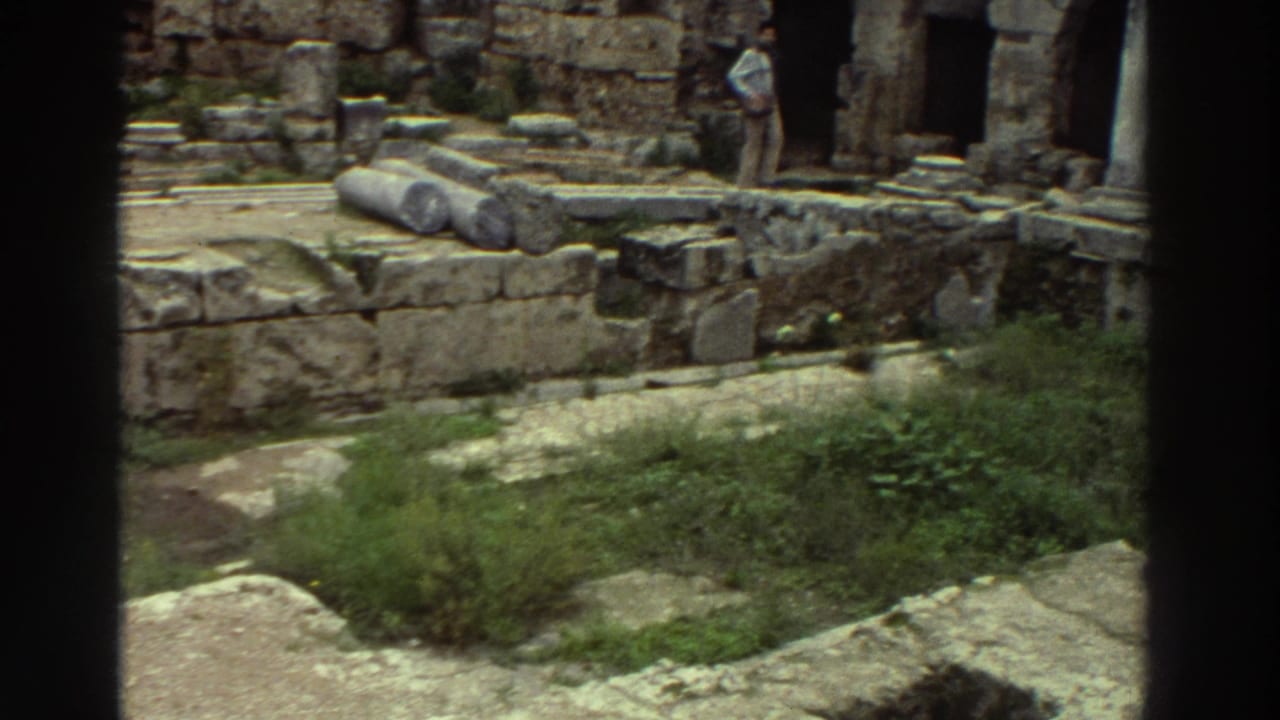 Wide view of ancient stone ruins with arched niches, fallen column drums, and green vegetation; tourists visible in background.