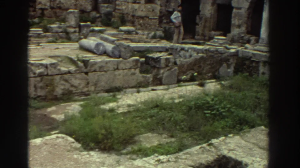 Wide shot of the Peirene Fountain ruins showing arched stone niches, fallen column drums, and overgrown vegetation, with tourists in background, Greece, c. 1976.