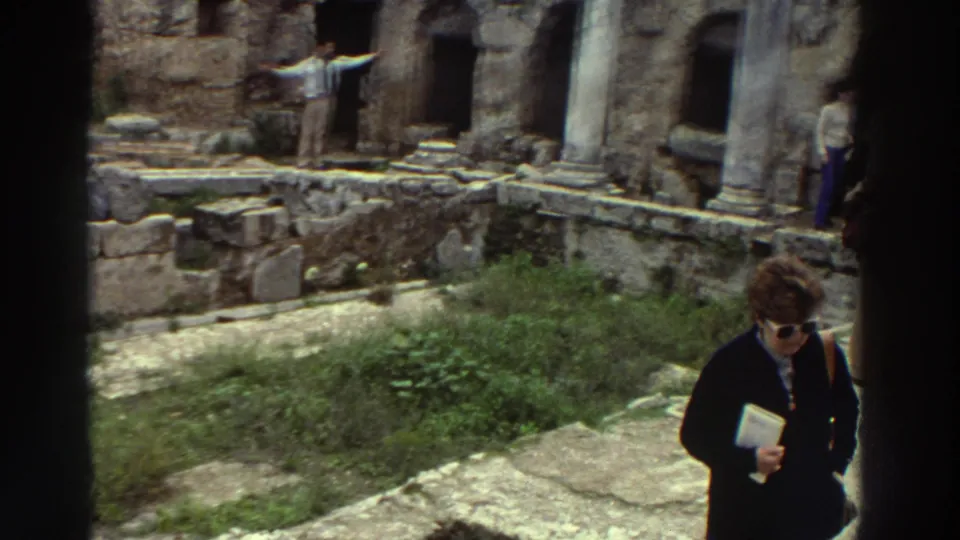 Female tourist in dark coat with sunglasses and guidebook walks through the Peirene Fountain site, arched niches and standing columns visible behind, Greece, c. 1976.