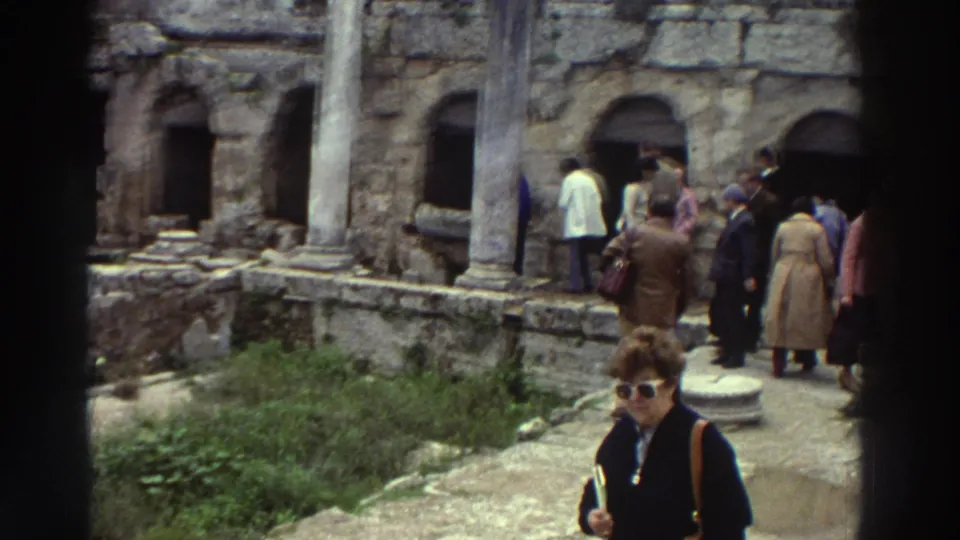 Several tourists explore the Peirene Fountain ruins; a woman with sunglasses and shoulder bag stands in foreground, stone arches and partial columns behind, Greece, c. 1976.