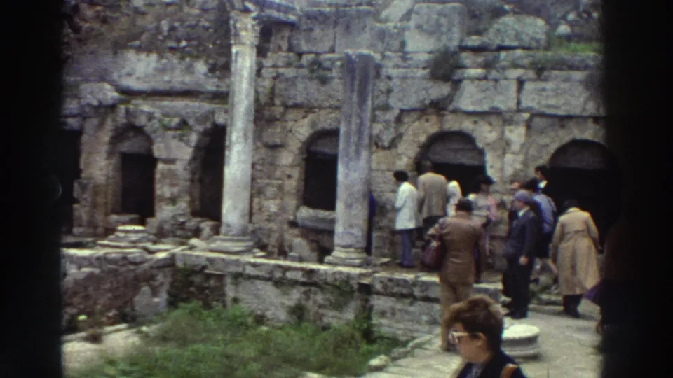 1970s tourists exploring a Greek classical ruin site, with arched stone niches, standing columns, and a woman in large-frame sunglasses close to the camera.
