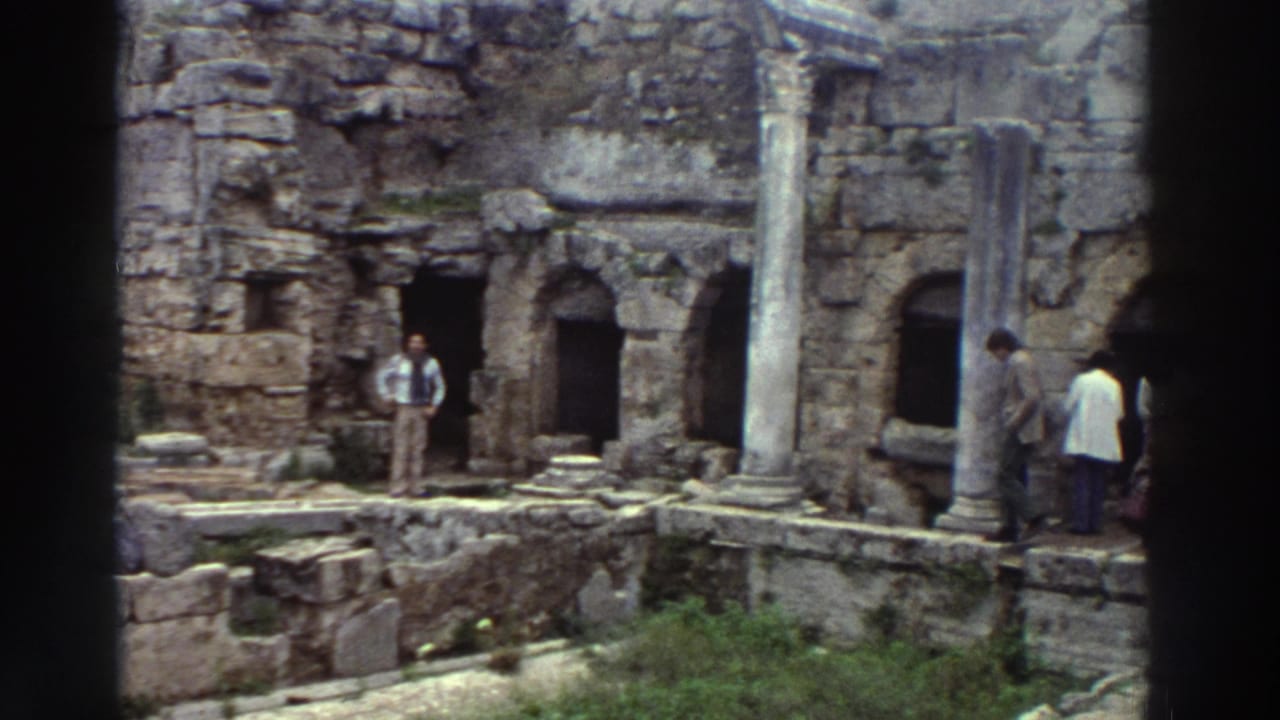 Two or three figures standing among ancient stone ruins with arched niches and columns; fewer visitors visible than in the preceding frame.