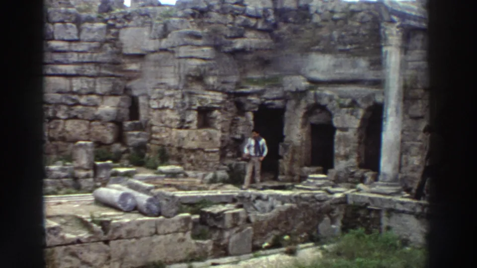 Wide shot of classical ruins in Greece, c. 1976, with fallen cylindrical column drums in the foreground, a standing column, and an arched niches wall behind distant figures.