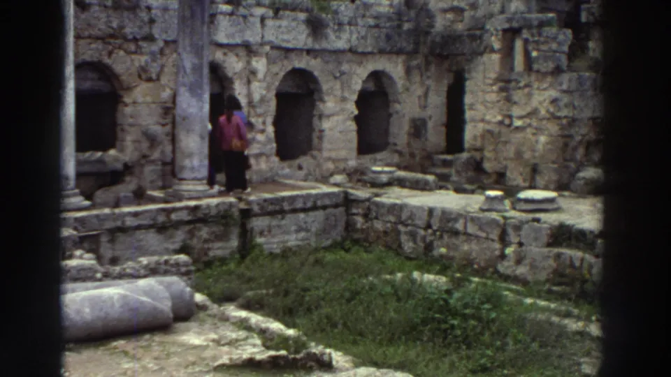 Two visitors in bright 1970s clothing pause beside columns at a classical Greek ruin site, c. 1976; fallen stone drums and grass visible in the foreground.