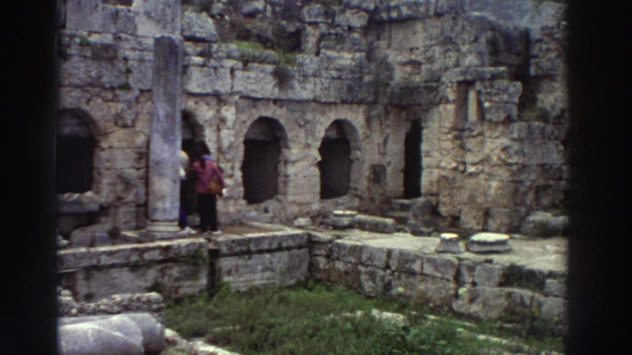 Two figures near ancient stone ruins with arched niches; one carries a brightly colored item; fallen column drums lie on the ground.