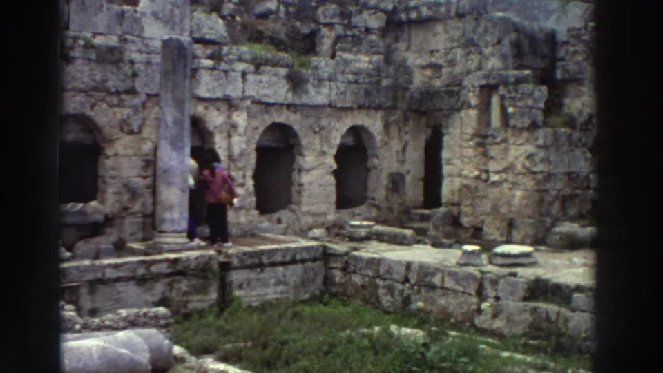 Two tourists near ancient ruins with arched niches and scattered column drums, one carrying a bright-colored item, Greece, 1976 8mm home movie.