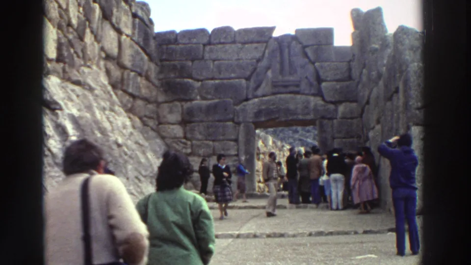 Crowd of 1970s tourists in winter coats gathered before what appears to be a monumental ancient stone gateway with cyclopean masonry, Greece, 1976.