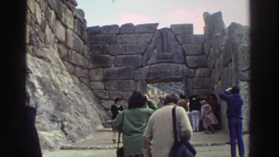 Tourists including a woman in a green coat view what appears to be a monumental cyclopean stone gateway, some photographing, Greece, 1976 8mm home movie.