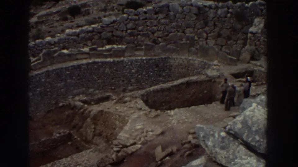 8mm wide shot of the Mycenae citadel excavation, Greece, c.1976, with small figures of tourists visible among the terraced stone ruins.