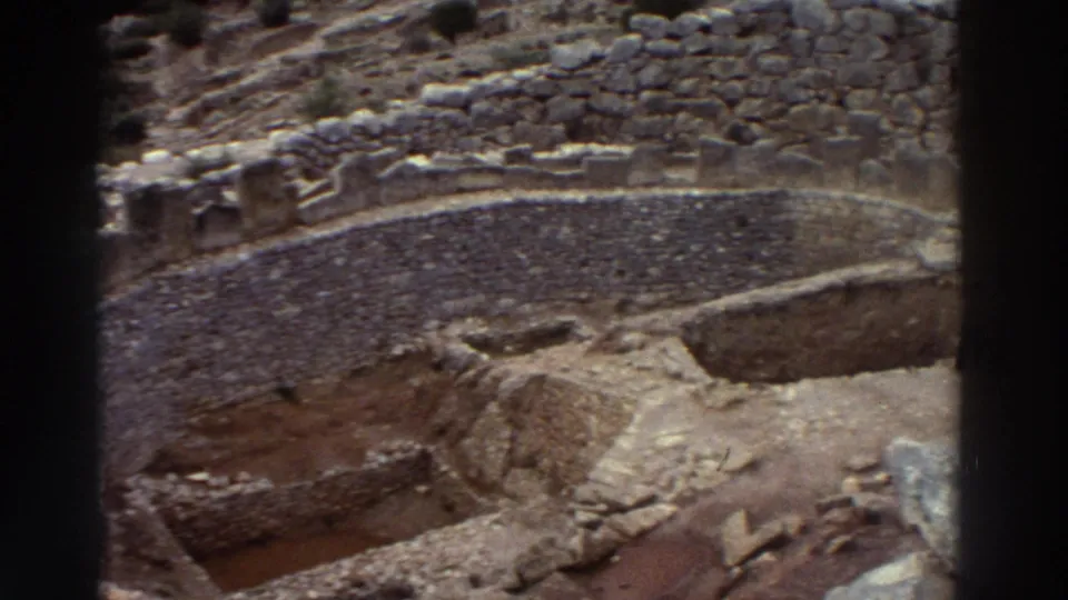 8mm wide-angle view of Mycenae citadel excavation terraces, Greece, c.1976, emphasizing the scale of ancient stone construction against the rocky landscape.