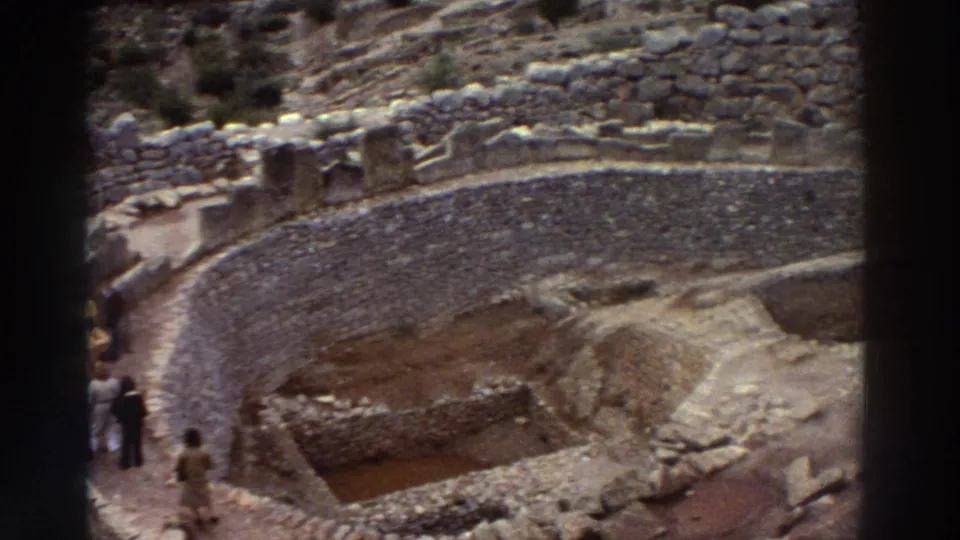 8mm footage from Greece, 1976: elevated view of dry-stone curved ruins on a hillside with one or two small figures visible at lower left.