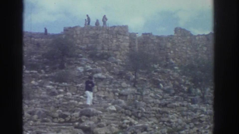 8mm home movie wide shot at a Greek ancient site, 1976: a tourist in white trousers walks the rocky slope while others stand atop ruined walls