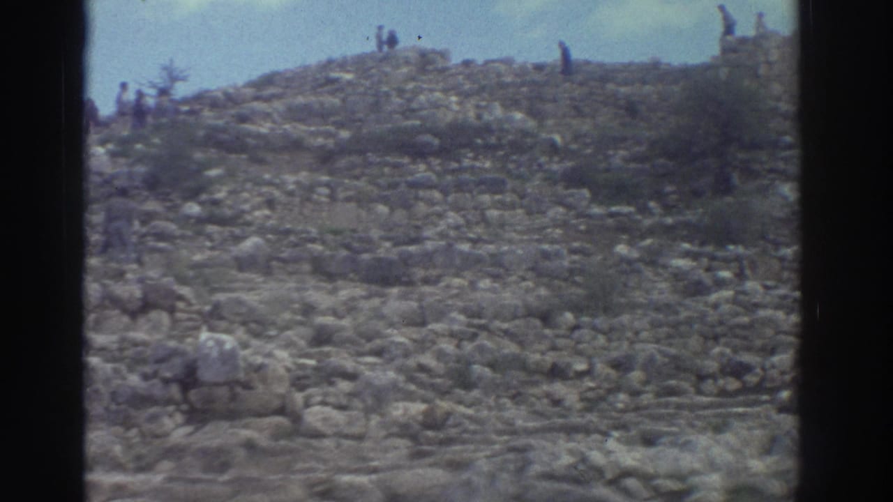 Rocky hillside covered in ancient stone ruins with scattered visitors climbing the slope