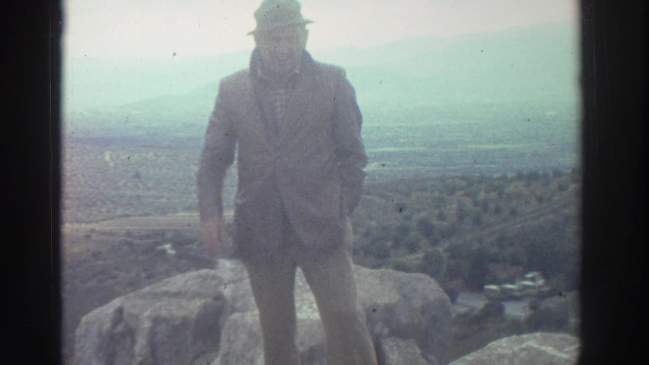 Man in suit and wide-brimmed hat standing on a rocky outcrop with a hazy valley landscape behind him.