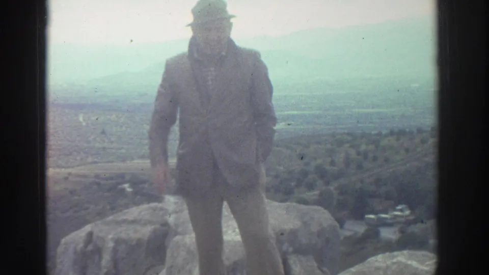 Male tourist in suit and wide-brimmed hat poses on rocky terrain at a Greek archaeological site, circa 1976, with a broad hazy valley visible in the background.