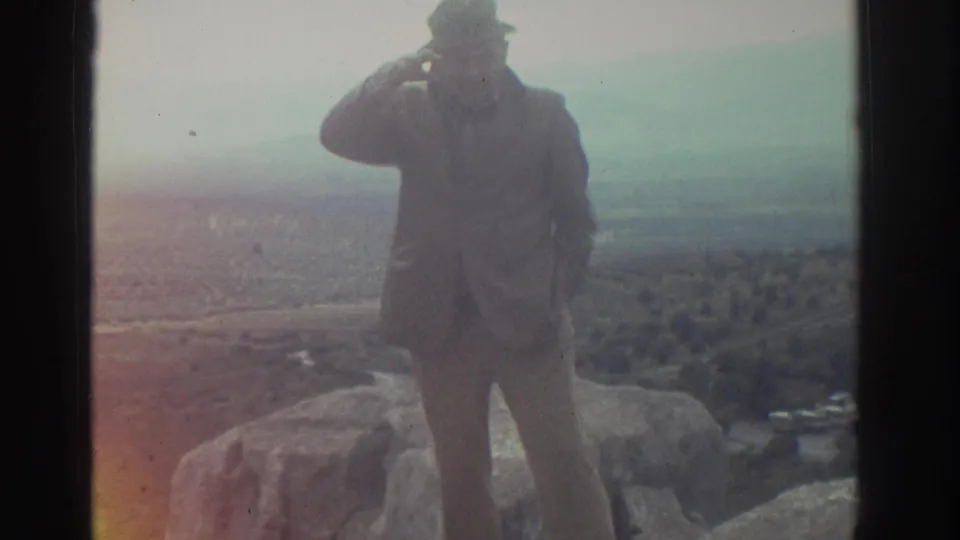 Tourist seen from behind stands on rocky outcrops at a Greek ruins site circa 1976, surveying a broad hazy valley; color fringing visible at the frame edge.