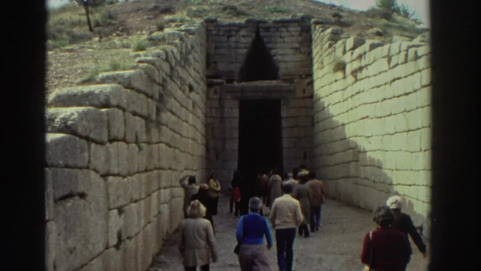 Group of tourists in 1970s clothing walk a stone dromos toward what appears to be a corbelled tholos tomb entrance at a Greek archaeological site, circa 1976.