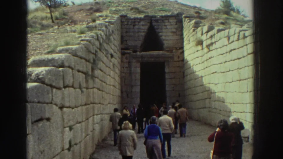 Tourists in 1970s clothing walk toward the entrance of what appears to be a Mycenaean tholos tomb in Greece, c. 1976, viewed from the dromos in 8mm home-movie footage.