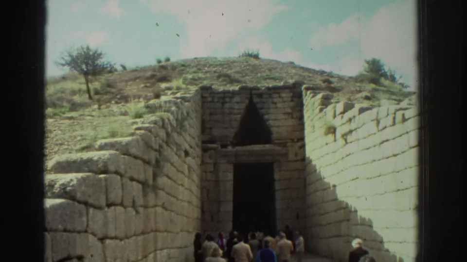 Angled view of cyclopean stone walls converging toward the entrance of a Mycenaean tholos tomb in Greece, c. 1976, with tourists near the doorway and a bare tree at upper left.