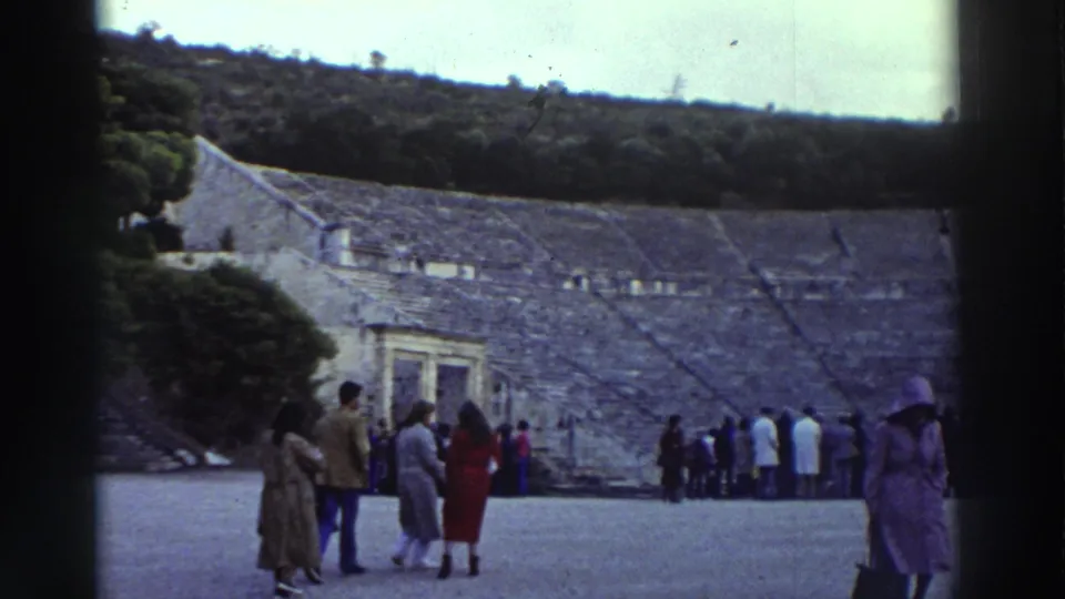 Tourists on the orchestra floor of a large ancient Greek theater with curved tiered stone seating, filmed in Greece c. 1976 on 8mm home-movie stock.