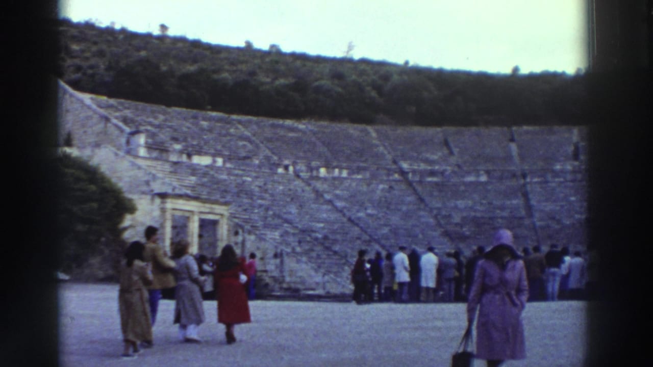 8mm home movie frame, circa 1976, Greece: tourists on the orchestra floor of a large ancient stone theater; woman in purple coat and hat foreground; stone seating tiers and wooded hill beyond.