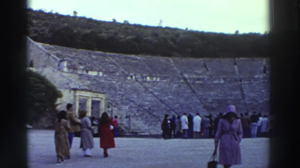 8mm home movie frame, circa 1976, Greece: tourists on the orchestra floor of a large ancient stone theater; woman in purple coat and hat foreground; stone seating tiers and wooded hill beyond.