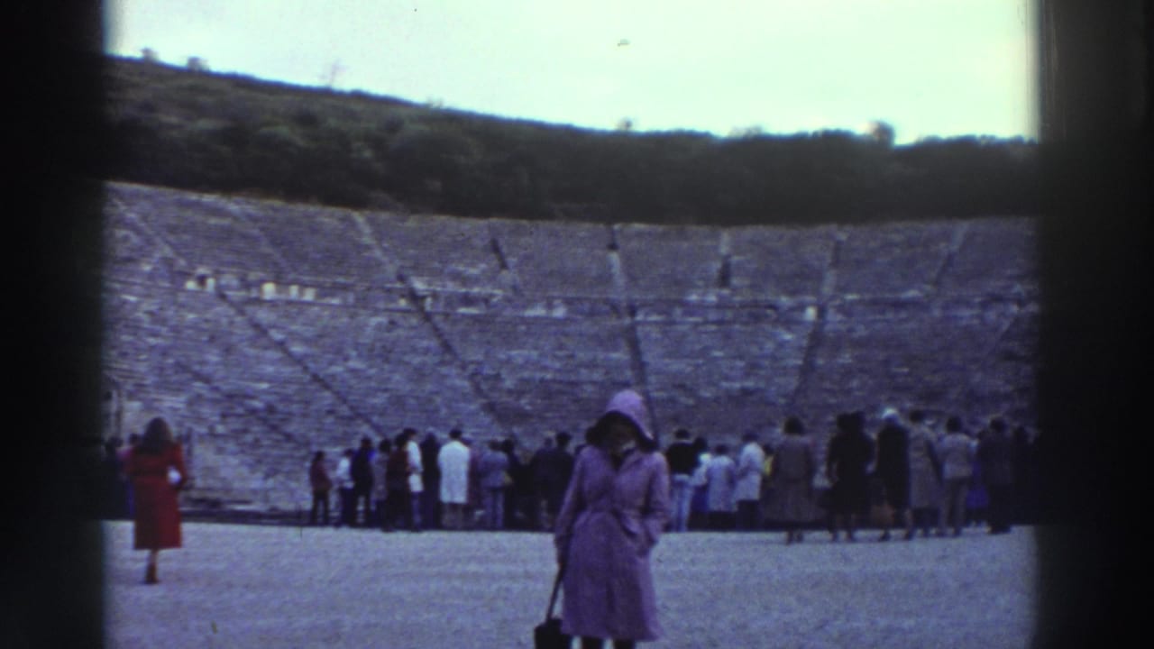 Wide view of a large ancient stone amphitheater; crowd of tourists gathered at the stage end; woman in purple coat stands in right foreground.