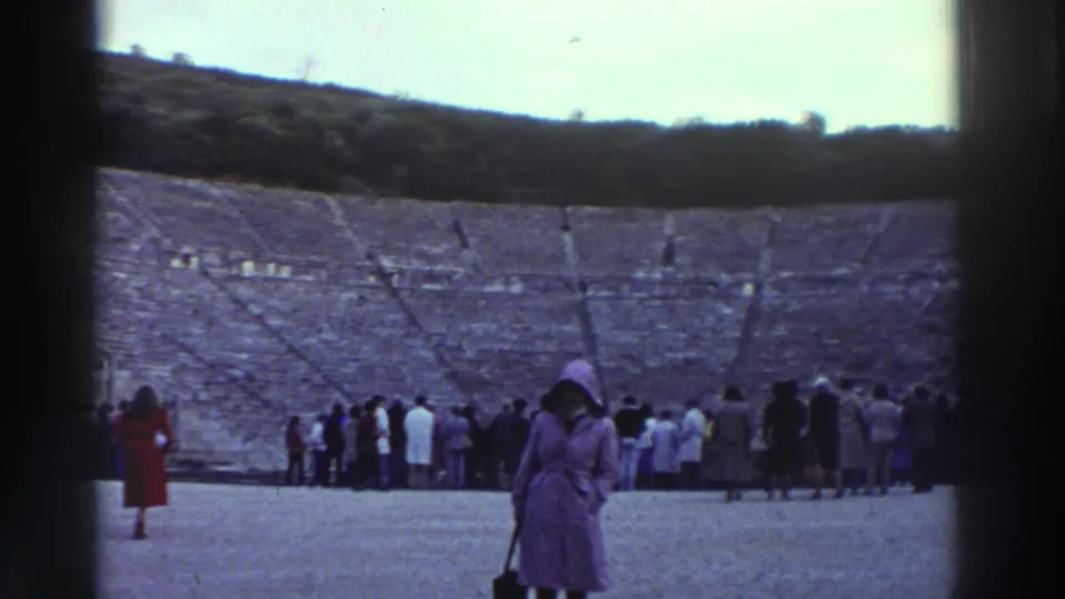 8mm home movie frame, circa 1976, Greece: wide view of ancient stone theater; large tourist crowd gathered near the stage; woman in purple coat and hat right foreground; stone tiers and hills beyond.
