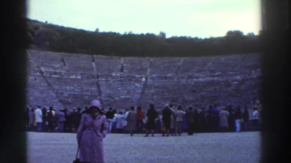8mm home movie frame, circa 1976, Greece: pan across ancient theater orchestra floor; wooden poles or posts midground; woman in purple coat foreground left; large tourist crowd at stage end; upper stone tiers above.