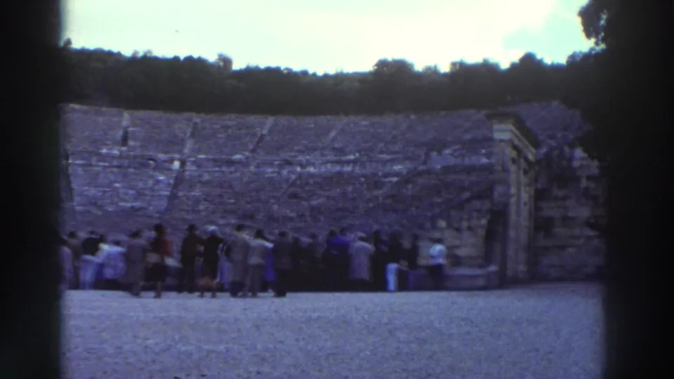 8mm home movie frame, circa 1976, Greece: end of pan showing stone tiered seating and small stone structure at right edge; crowd near stage end; woman in purple coat left; wooden poles visible on orchestra floor.