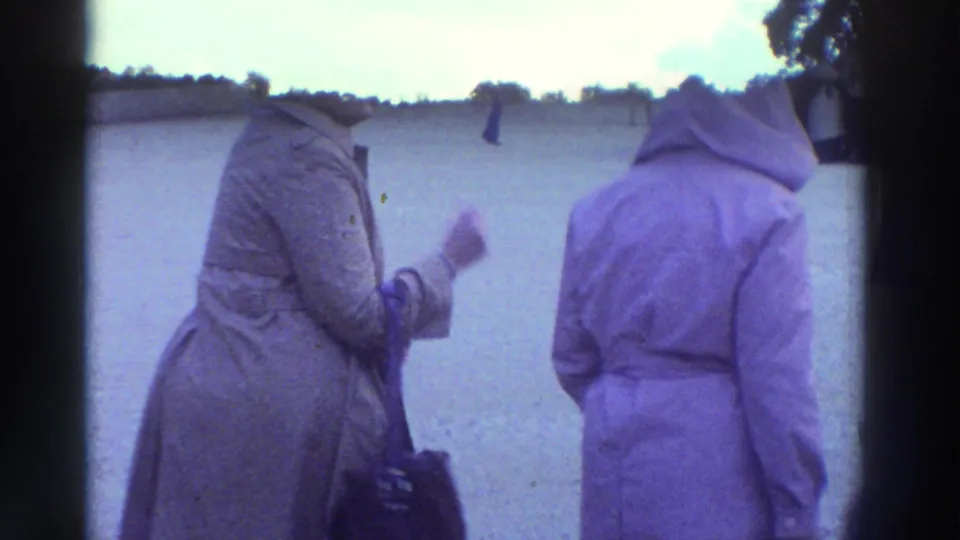 Two women in mid-1970s coats—one tan trench coat, one purple hooded coat—stand at what appears to be an ancient Greek archaeological site, circa 1976.