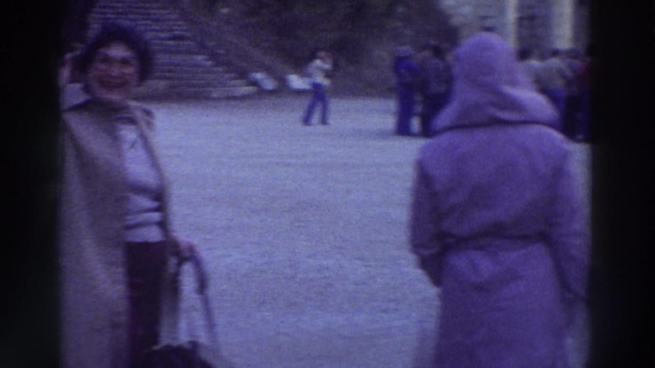 A smiling woman in a tan coat faces the camera; a second visitor in a purple hooded coat stands to her right.