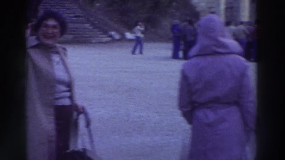 A smiling woman in a 1970s tan coat poses for the camera at an ancient Greek archaeological site in 1976, with other visitors visible in the paved area behind her.