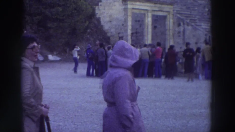 Tourists in 1970s clothing gather in the courtyard of an ancient Greek archaeological site in 1976; stone walls with arched niches are visible behind the crowd.