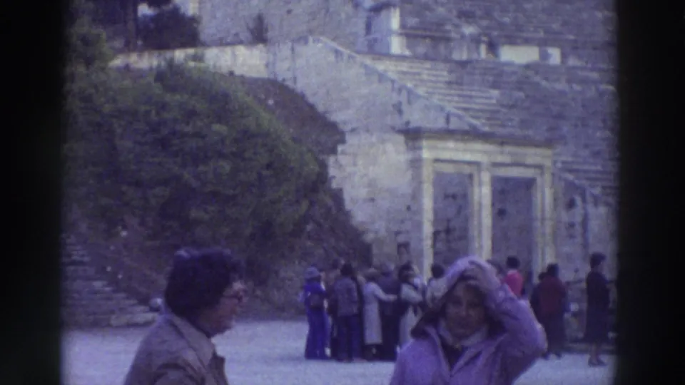 Tourists explore an ancient Greek site in 1976; stone walls with arched niches and tiered stone seating steps are visible in the background, consistent with an ancient theater.