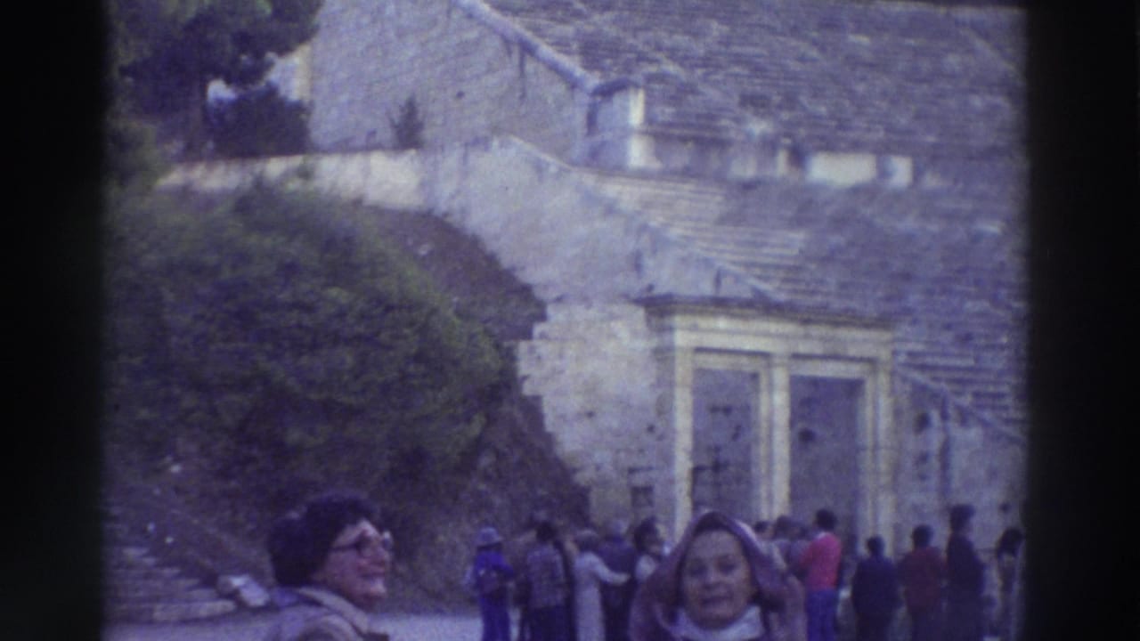 8mm home movie frame, circa 1976, showing tourists at an ancient Greek theater — woman with glasses in foreground, stone seating tiers and arched doorways rising behind.