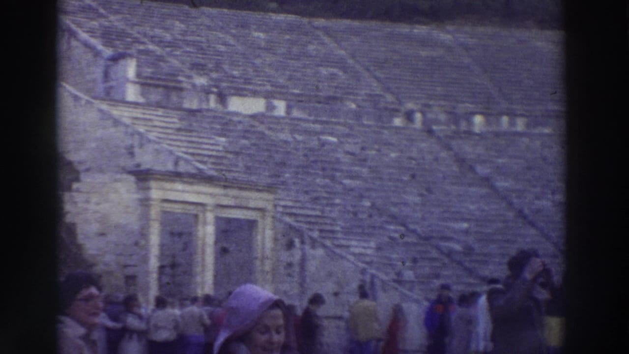 Tourists stand near the stone seating wall of an ancient theater; a woman in a purple hooded coat appears at right center.