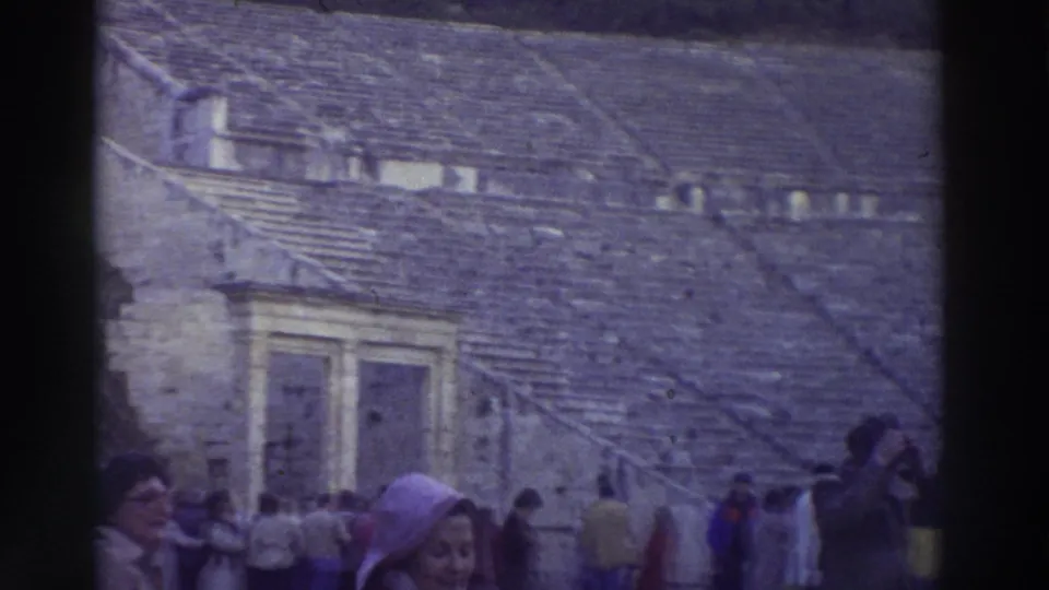 8mm frame from a 1976 Greek tour showing visitors in period coats at an ancient stone theater, woman in lavender hooded coat prominent at right.