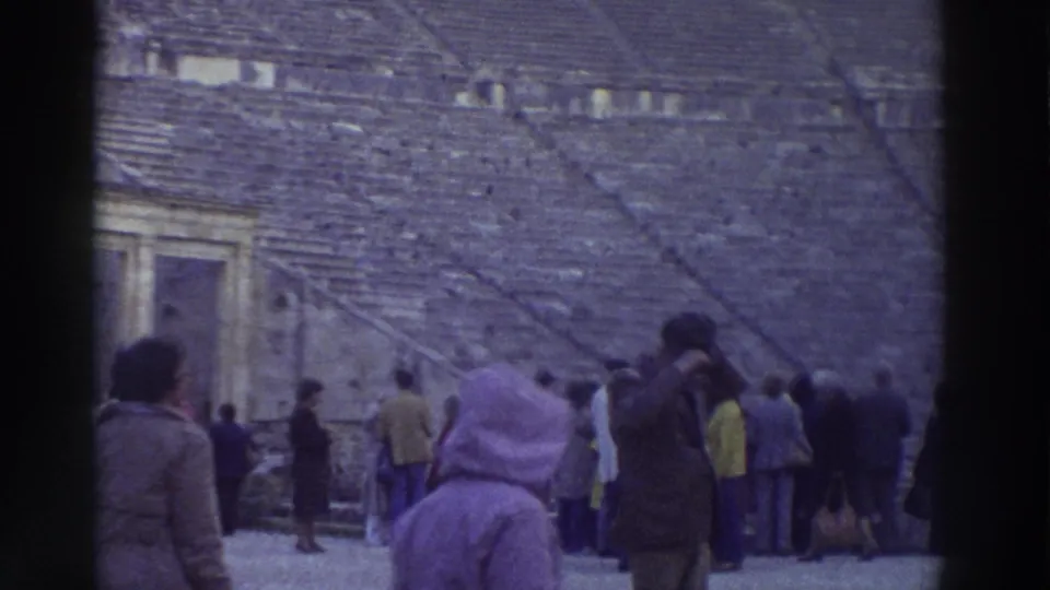 8mm home movie frame showing a woman in a lavender hooded coat crossing the orchestra area of an ancient Greek theater, c. 1976, with tourists clustered behind.