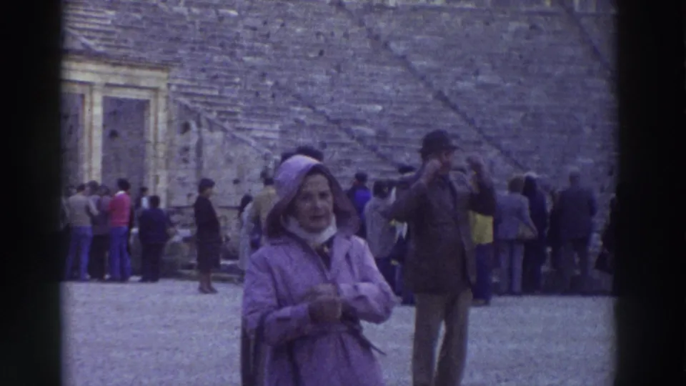 Close 8mm frame from Greece, c. 1976 — older woman in lavender hooded coat faces toward camera at an ancient theater, tourists and stone seating visible behind.